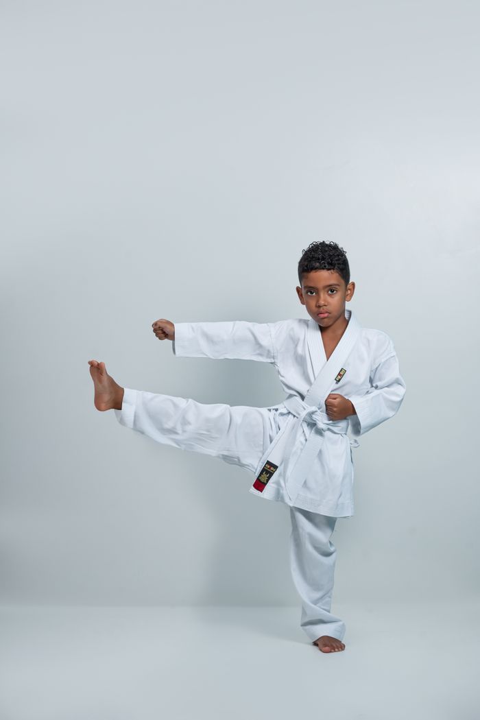 heros-img A young boy demonstrates a karate kick in a studio setting, wearing a white kimono.